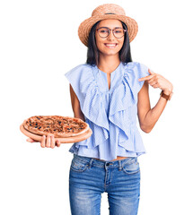 Young beautiful latin girl wearing summer hat and glasses holding pizza pointing finger to one self...