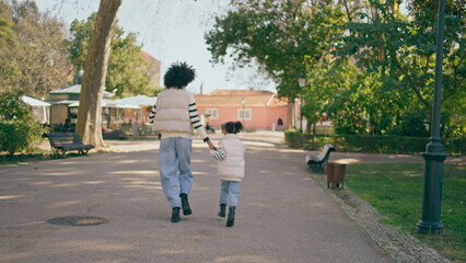 Fototapeta na wymiar African mother daughter running down park road sunny day. Back view happy family
