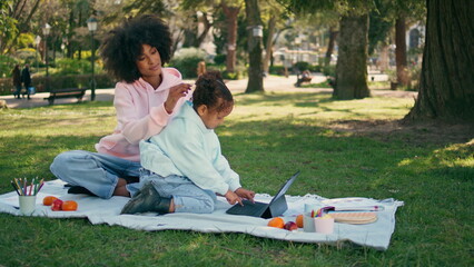Mother watching daughter tablet playing on picnic. African woman admiring girl 