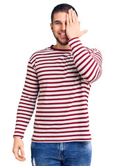 Young handsome man wearing striped sweater covering one eye with hand, confident smile on face and surprise emotion.