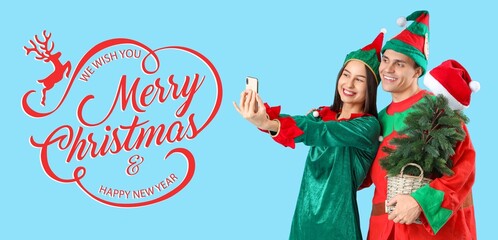 Happy couple in elf's costumes, with fir tree and mobile phone taking selfie on blue background....