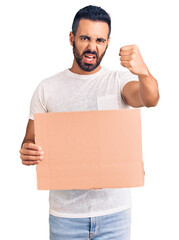 Young hispanic man holding banner cardboard annoyed and frustrated shouting with anger, yelling crazy with anger and hand raised