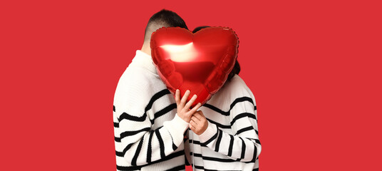 Loving young couple hiding faces behind heart-shaped balloon on red background. Valentine's Day...