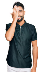 Young man with beard wearing sportswear covering one eye with hand, confident smile on face and surprise emotion.