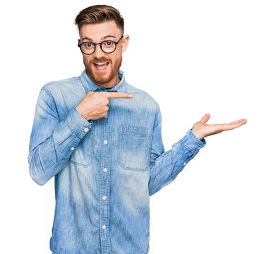 Young redhead man wearing casual denim shirt amazed and smiling to the camera while presenting with hand and pointing with finger.