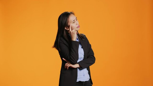 Thoughtful businesswoman posing over orange backdrop, thinking about new ideas and being in doubt. Clueless uncertain person brainstorming solutions, acting focused in front of camera.