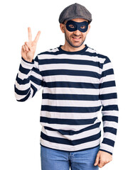 Young handsome man wearing burglar mask smiling looking to the camera showing fingers doing victory sign. number two.