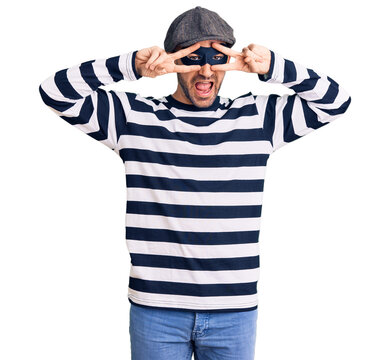 Young handsome man wearing burglar mask doing peace symbol with fingers over face, smiling cheerful showing victory