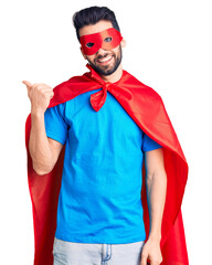 Young handsome man with beard wearing super hero costume smiling with happy face looking and pointing to the side with thumb up.