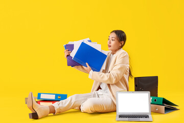 Young Asian businesswoman with document folders and laptop on yellow background