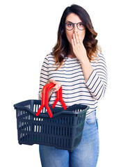 Beautiful young brunette woman holding supermarket shopping basket covering mouth with hand, shocked and afraid for mistake. surprised expression