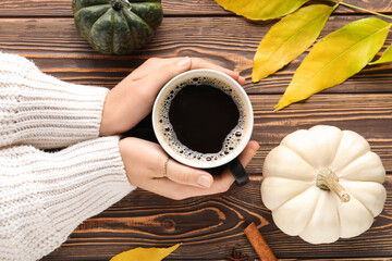 Female hands with cup of aromatic coffee, pumpkins and fallen leaves on wooden background, closeup