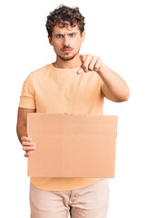 Young handsome man with curly hair holding blank cardboard empty banner pointing with finger to the camera and to you, confident gesture looking serious