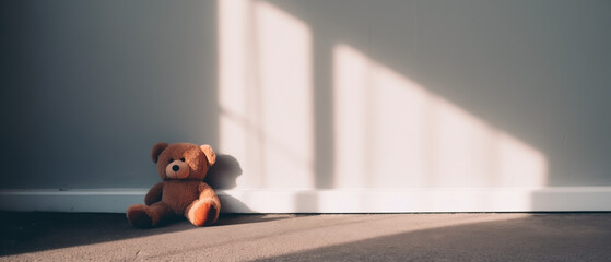 Lonely forgotten stuffed bear toy sitting against an empty room with morning sunlight and shadows against the wall, sad broken spirit waiting for someone to pick him up and take to a new home. - obrazy, fototapety, plakaty