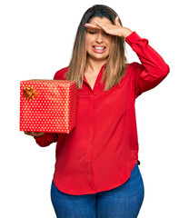 Beautiful brunette woman holding gift stressed and frustrated with hand on head, surprised and angry face