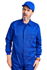 Middle age handsome man wearing mechanic uniform in shock face, looking skeptical and sarcastic, surprised with open mouth