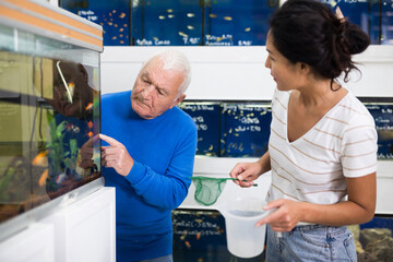 Asian woman helping old man to choose fish for his aquarium. He's pointing with finger on fish he...