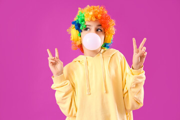 Trendy girl in clown costume blowing bubble gum while showing victory gestures on purple background