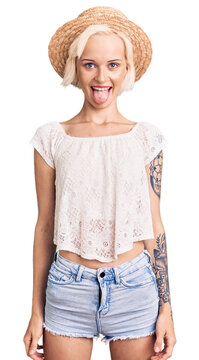 Young blonde woman with tattoo wearing summer hat sticking tongue out happy with funny expression. emotion concept.