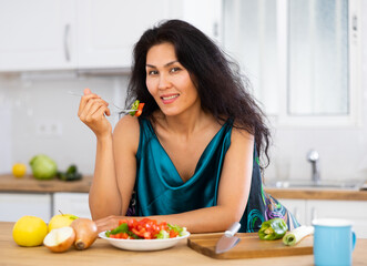 Obraz na płótnie Canvas Portrait of positive asian woman in nightie eating salad in kitchen at home. Woman has cooked fresh vegetable salad.
