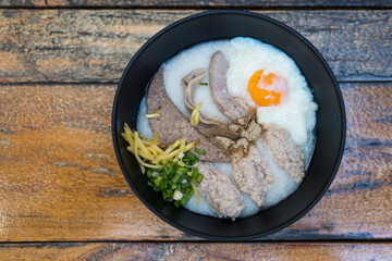 Close up of congee with soft boiled egg and pork liver, pork intestines, ginger sliced, spring onion sliced, coriander sliced in black bowl on wooden table.