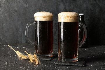 Mugs of cold dark beer with wheat on table against black grunge background