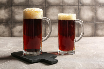 Mugs of cold dark beer on table against grey tile background