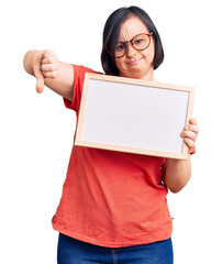 Brunette woman with down syndrome holding empty white chalkboard with angry face, negative sign showing dislike with thumbs down, rejection concept