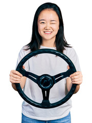 Young chinese woman holding steering wheel winking looking at the camera with sexy expression,...