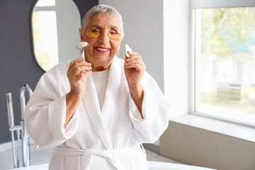 Senior woman with under-eye patches and facial massage tools in bathroom