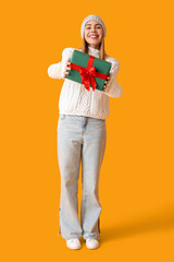 Happy young woman in winter clothes with Christmas gift on yellow background
