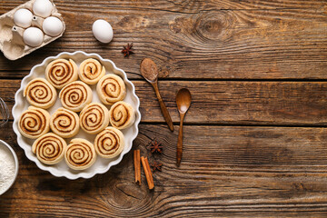 Baking dish with uncooked cinnamon rolls and ingredients on wooden background