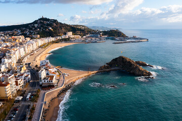 Fototapeta premium Scenic drone view of coastal Catalan town of Blanes overlooking white residential buildings and Sa Palomera Rock on background of San Juan hill with ancient castle on sunny day, Barcelona, Spain ..
