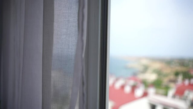 Sea view through the window with white transparent curtains slowly swaying in the wind. Resort, apartment with sea view. Leisure, relax, beauty, travel.