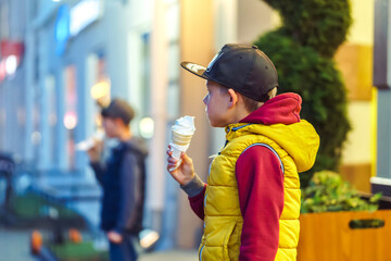 Boys in cap and vest eats tasty ice cream in evening city. Kid holds healthy cold dessert walking...