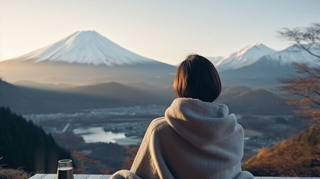 Back view over-the-shoulder Shot of the lady wearing cold weather clothes sitting by the Fuji mountains view. Woman relaxes by mountain view with a cup of hot drink