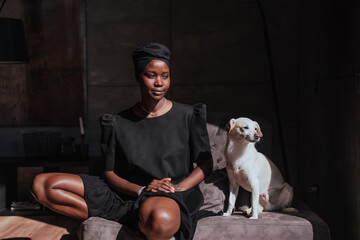 Young African girl in black turban and black traditional clothes sitting on couch with white dog...