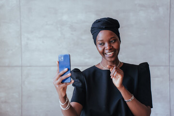 Joyful African girl in black turban talks by phone makes video call against marble wall with copy...