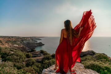 Woman sunset sea red dress, back view a happy beautiful sensual woman in a red long dress posing on a rock high above the sea on sunset.