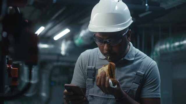 Professional African American engineer eats sandwich and uses phone surfing Internet. Heavy industry worker in safety uniform, goggles, protective hard hat having break at modern manufacturing plant.