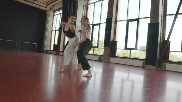 Long shot of two Caucasian female dancers demonstrating acrobatic elements while dancing contemporary on red parquet floor of big loft style dance studio with panoramic windows