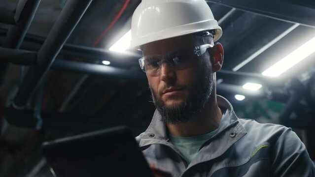 Close up of professional Caucasian engineer using digital tablet computer on industrial manufacturing factory. Heavy industry specialist in safety uniform, goggles and hard hat works on modern plant.
