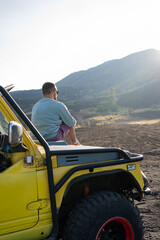 man enjoying beautiful rocky landscapes, sitting on the car hood on the roadside. Traveling on the volcanic valley in Bali.