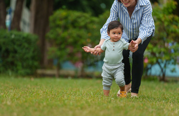 infant baby learn walking first step on grass field with mother holding hand helping in park