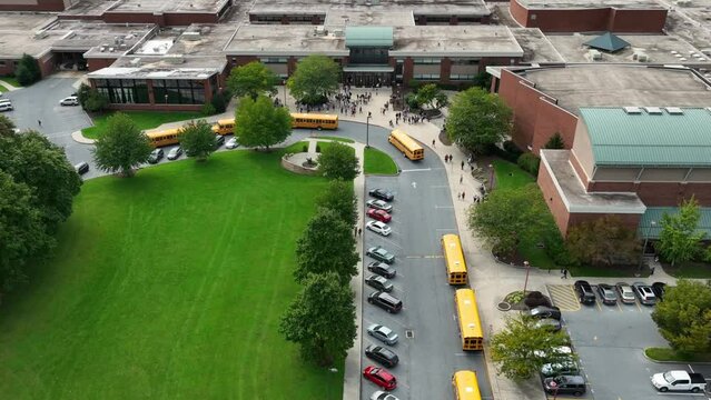 Students at public school campus buildings as school buses arrive to transport learners. Diversity DEI theme in American education. Aerial.