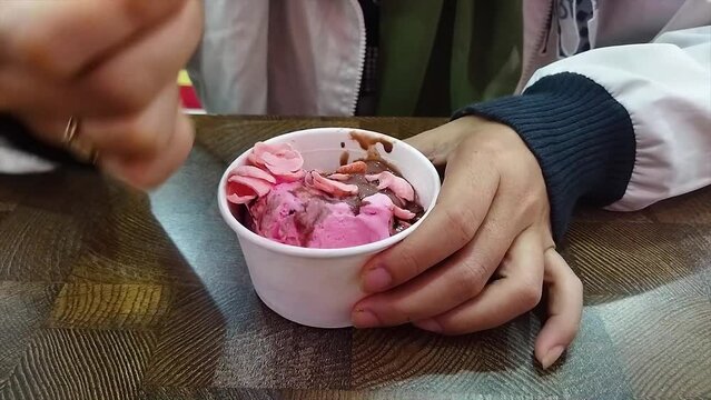 Young woman's hands eating ice cream in a white cup at the table while sitting in an ice cream shop. Ice cream in a cup with strawberry, chocolate and Coco Crunch topping