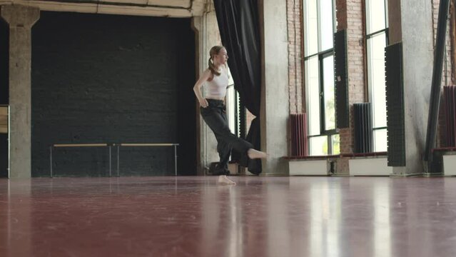 Long shot of young well-trained Caucasian woman in comfortable relaxed sportswear dancing contemporary freestyle on red parquet in loft style dance class
