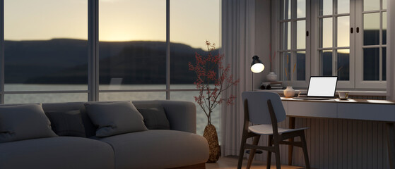 A modern living room in the evening with a cosy couch near by the window and a laptop on a table.