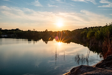 This image captures the serene atmosphere of a quarry lake at sunset. The sun hovers just above the treeline, casting a soft glow over the still water. The lake, flanked by rocky edges and sparse - obrazy, fototapety, plakaty