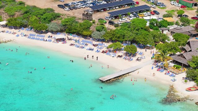 Drone orbit around pier at Playa Porto Mari as tourists swim and walk on white sand beach